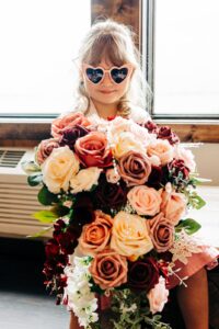 Flower girl holding bride's bouquet and wearing pink heart sunglasses before wedding