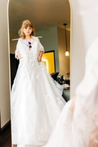 Bride's daughter holding mother's wedding dress up to herself in the mirror