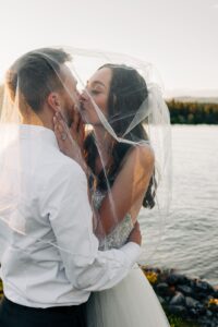 Bride and groom kissing under wedding veil on shore of Flathead Lake