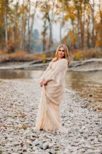 Pregnant woman in long dress standing on bank of Clark Fork River at dusk