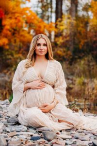 Pregnant woman sitting on river rocks with orange foliage in background