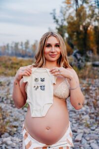 Pregnant woman holding up baby onsie at Kelly Island in Missoula, MT