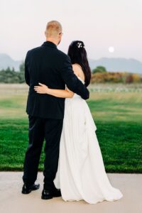 Portrait of bride and groom standing arm-in-arm facing supermoon rising over Swan Mountains at Snowline Acres