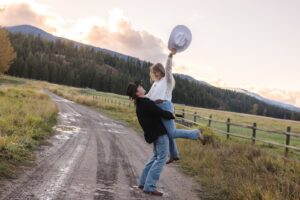 Man lifting woman up on dirt road with mountains in background at sunset