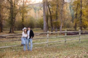 Engaged couple posing at wood fence with fall foliage in in background